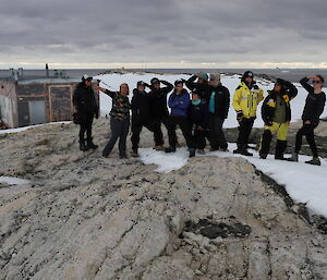 Group of 11 women standing mid picture on snow covered rocks, with wood field hut left of photo and sea and grey sky in background.