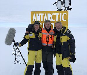 Three people on ice standing in front of yellow sign reading Antarctic Circle. Lady on left holding a large microphone, man in centre has arms over other two people and is wearing high vis orange jacket, man on right is Sam Neill. All wearing yellow and black AAD antarctic gear.