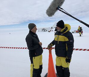 Two men talking, one on right is Sam Neill, wearing black and yellow AAD antarctic gear, striped tape at waist height behind, orange witches-hat on ground between. Large microphone in foreground, top centre. White plane on ice runway behind in distance.