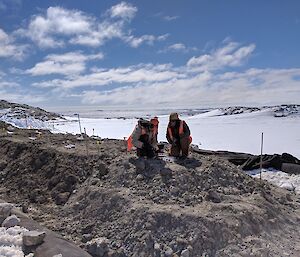Two expeditioners in winter clothing and high-vis vests working on soil mound, with icy landscape behind and blue sky with white cumulous clouds above.