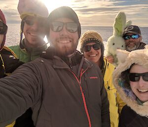 Team selfie of six people with sun and sea behind. All in assorted winter gear, in particular woman on right wearing a bear spirit hood with ears and man at back left holding a green toy bunny.