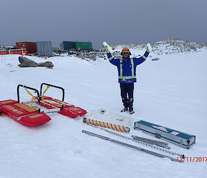 Ice coring equipment laid out on ice, Man in blue jacket with orange hard hat holding up two ice core samples, one in each hand. In the background Casey buildings in red and green.