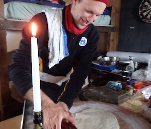 Man wearing red beanie with tea-towel over shoulder, rolling out a round of pizza dough with a wine bottle. Inside field hut with bunk bed behind and lit candle in wine bottle at front.