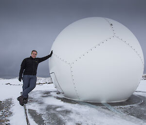 Man in black jumber and grey pants on left, leaning against a large white dome in centre picture. On icy concrete ground with grey sky overhead.