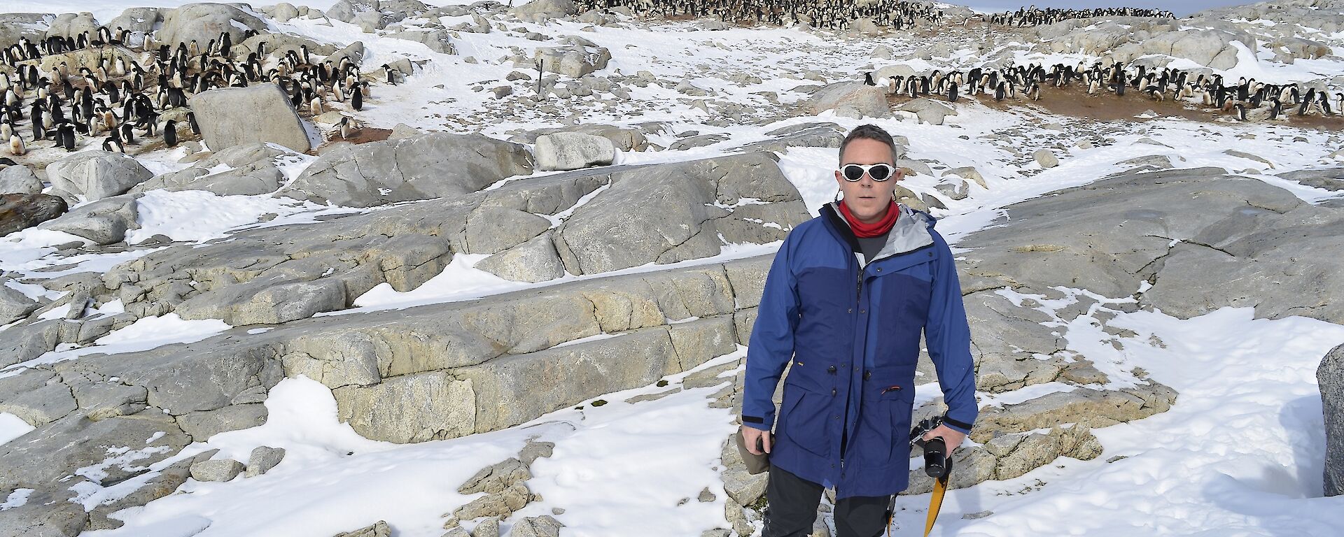 Blue in winter gear, black pants, blue jackets, white rimmed sun glasses, Standing on snow covered rocks with groups of penguins in the background.