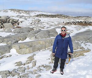 Blue in winter gear, black pants, blue jackets, white rimmed sun glasses, Standing on snow covered rocks with groups of penguins in the background.