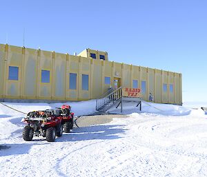 Mid picture, large yellow building with stairs going up to frond door in middle. Sign on stairs reads RADIO VNJ. Foreground is snow covered ground with two red quad bikes parked in front. Bright blue sky above.