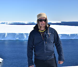 Man in blue outdoor clothing and sheeps wool hat standing centre photo with blue sea and glacier tongue in background. Blue sky above.