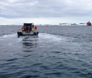 Foreground to mid horizen, blue seas. On horizen in distance the Aurora Australis, large orange ship. Mid picture, grey barge moving towards ship rolling out hose into water.