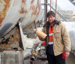 Left — large fuel tank in silver and rust. Right — man in high vis shirt and beige winter jacket holding a foam fuel pig which is torpedoe shaped approximately 5 inches diameter and 10 inches long, flat one end pointy the other.