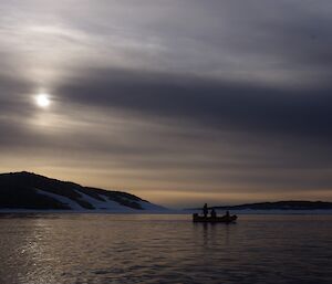 Dark gloomy sky with sun trying to peak through clouds. Foreground calm dark seas, mid picture rocky snow covered shoreline. Sillouted boat with three people mid picture.
