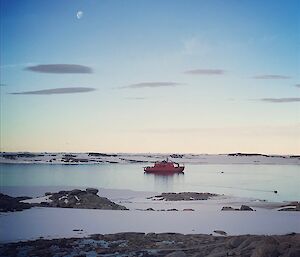 Aurora Australis, large red ship in centre of Bay, Shoreline is rocks and snow. Blue sky tinged with orange and yellow as if at sunset, small white 3/4 moon in top left.