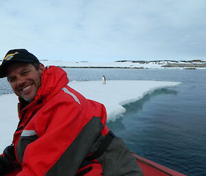 Man centre front in red and black dry suit and black ball cap is smiling, In background is large flat piece of ice-flow with a penguin standing on it off in the distance.