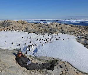 Women in black outdoor gear reclining on a rock in foreground. Middle of picture a large group of penguins on a patch of snow. Backgroun, rocky ground then sea and blue sky.