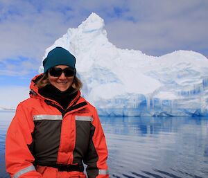 Woman in red and black dry suit with blue beanie and black sun glasses in front left of picture. Large iceberg in background.