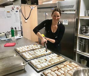 Kitchen setting, Front to left back long steel work bench with four trays of small rectangular lemon meringue tarts. Woman in black chefs uniform in centre back holding a torch to toast the top of the meringues.