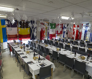 Dining room with five long tables with chairs. Paper snowflakes hanging from the roof and country’s flags hanging around the walls.