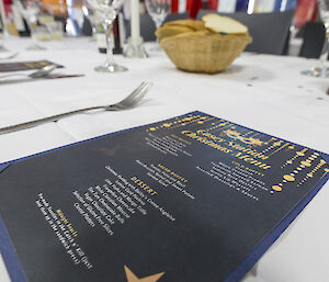 Menu for dinner on the table with cutlery either side. Menu is black with gold decorations and white writting.