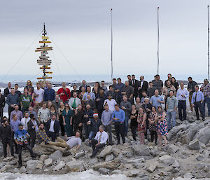 A large group (approximately 80) of people standing on rocky outcrop infront of casey sign and four flag poles. Newcomb Bay behind with overcast skies.