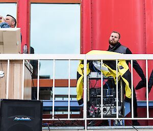 On balcony of red shed, On man on left behind DJ desk and two men on right dancing