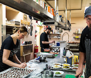 To left female chef putting finishing touches to christmas chocolates on a tray. Front right chef wearing striped apron and blue ball cap at chopping board. Back centre chef in black working at the kitchen bench.