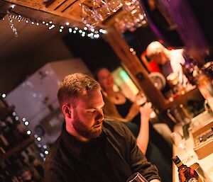Man in black shirt with cocktail mixer centre front, bar scene behind