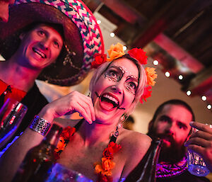 Centre picture woman with face pointed as “day of dead’ skull with flowers in hair, laughing. To left one man with sombrero. To right a third man with colourful scarf raising glass.