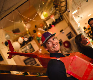 Man wearing cap with helicopter propellor on head and cardboard costume of a helicopter in red over shoulders. Behind is a staircase and in the ceiling are balloons and christmas decorations