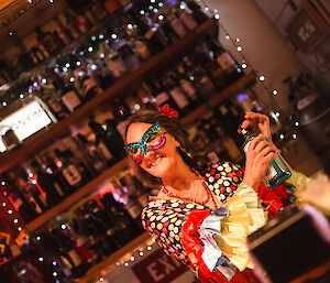 Lady wearing bright spanish flamenco outfit with butterfly mask over eyes, standing in bar scene holding a bottle