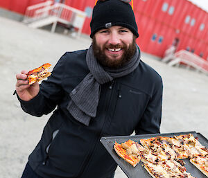 Man in black with grey scarf and black beanie, holding a tray of pizza in left hand and eating a slice from right hand.