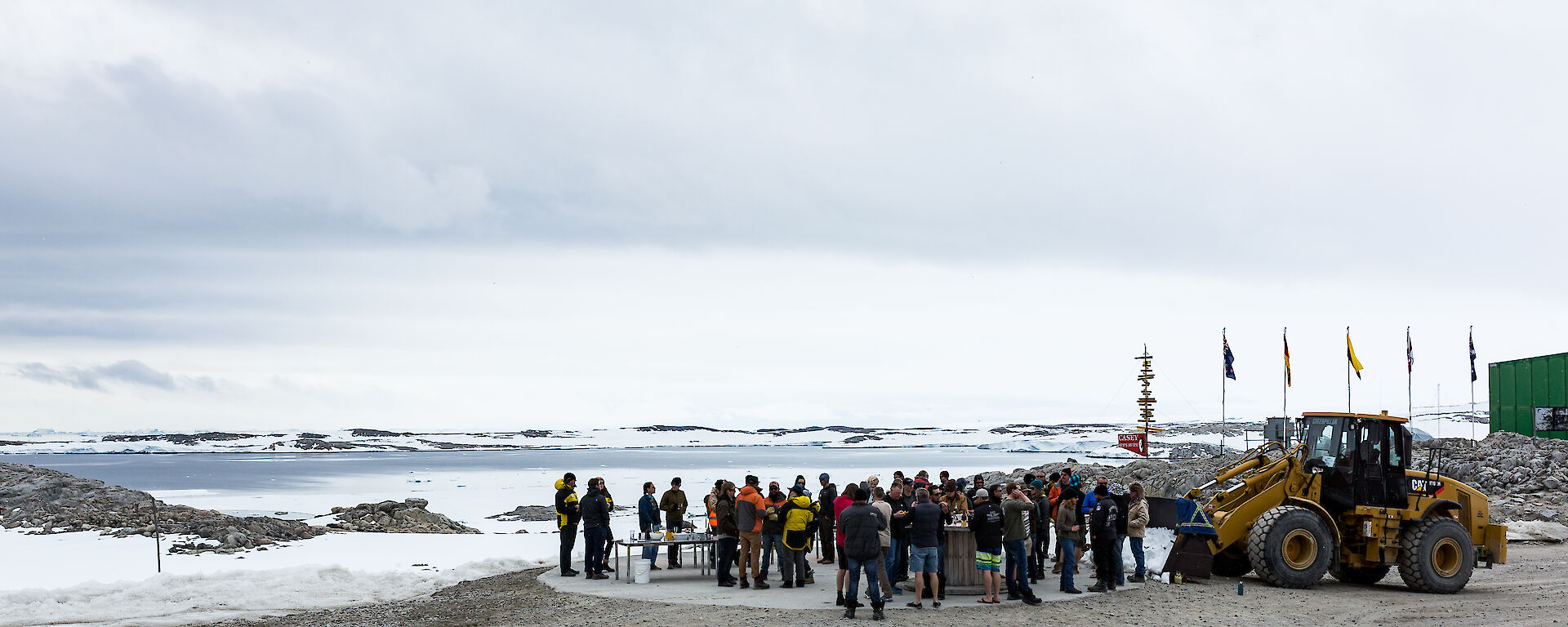 Foreground large group of people standing beside a bulldozer, mid picture snow and rocks with five flag poles and casey sign, distance the bay with cloudy skies above.