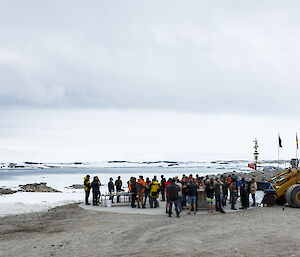 Foreground large group of people standing beside a bulldozer, mid picture snow and rocks with five flag poles and casey sign, distance the bay with cloudy skies above.