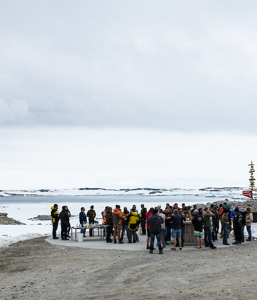 Foreground large group of people standing beside a bulldozer, mid picture snow and rocks with five flag poles and casey sign, distance the bay with cloudy skies above.