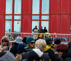 Crowd of poeple in foreground, in background three people on balcony of the red shed.