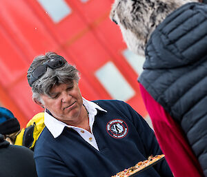 Man in blue rugby top holding a tray of pizza, in amongst a group of people. Bright red building in background.