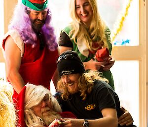 Man sitting on santas knee receiving gift wrapped in red paper, behind is a elf with purple hair and red and green stripped cap, and an elf in green dress wearing green reindeer antlers