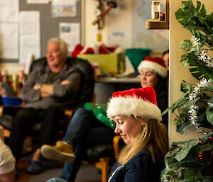 Woman in santa hat in centre of picture, other people sitting around in lounge area.