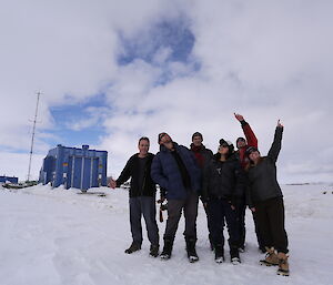Group of six people looking and pointing into the sky at front right on snow covered ground. In left background a blue square building — the hydrogen balloon shed at Casey
