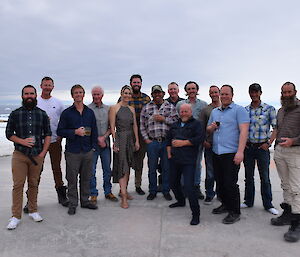 A group of 14 people in a group standing on concrete helipad with ice and rock cover ground leading to bay and grey sky