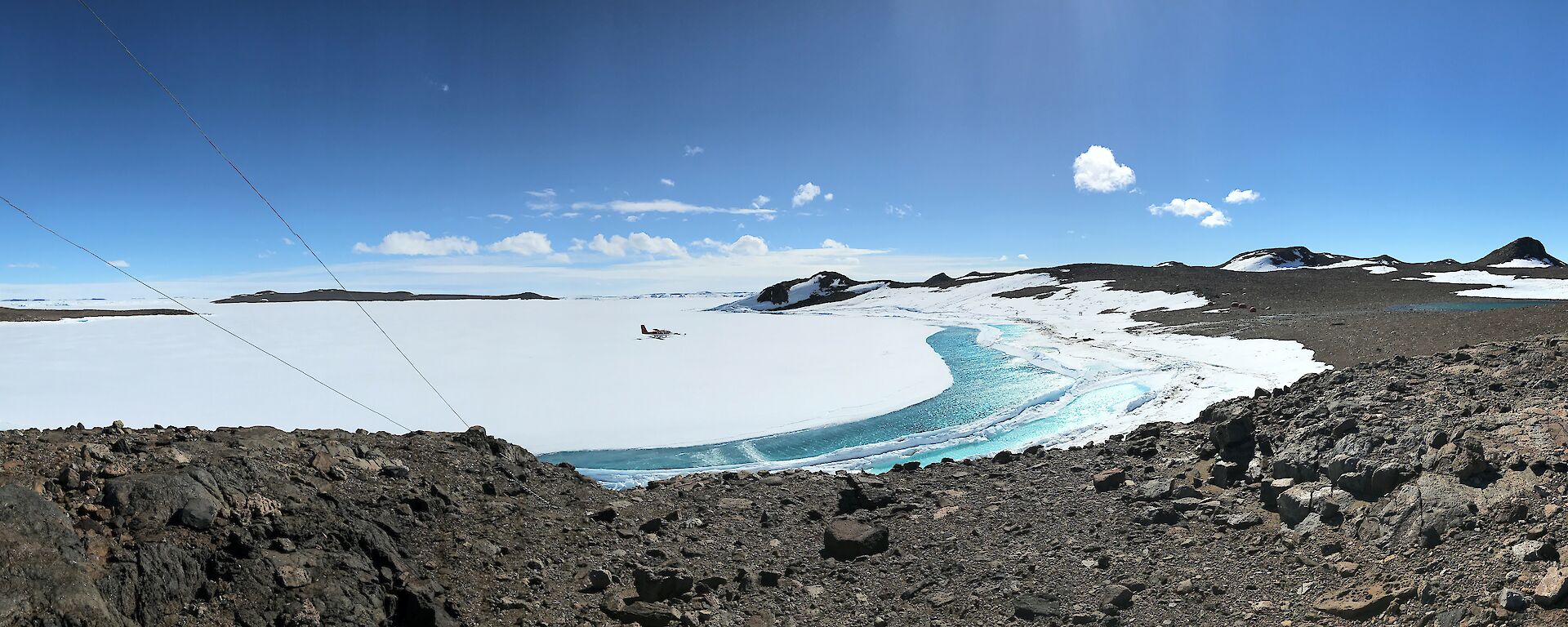 Panaroma shot showing AWS instrument to left of photo and then view from rocky hill across ice covered bay to rocky shoreline. Melt pools of aqua water between sea-ice and shore in centre of photo. Blue sky above.