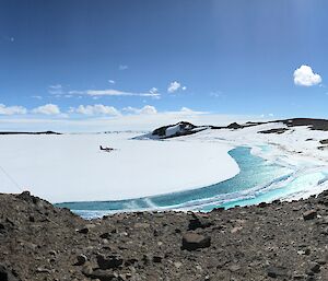 Panaroma shot showing AWS instrument to left of photo and then view from rocky hill across ice covered bay to rocky shoreline. Melt pools of aqua water between sea-ice and shore in centre of photo. Blue sky above.