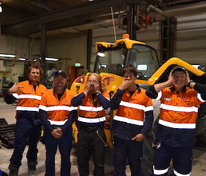 Five men in high vis clothing standing in front of yellow bulldozer in workshop, left to right, one pointing right, one covering crutch, one covering eyes, one covering mouth, one covering ears