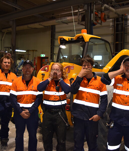 Five men in high vis clothing standing in front of yellow bulldozer in workshop, left to right, one pointing right, one covering crutch, one covering eyes, one covering mouth, one covering ears