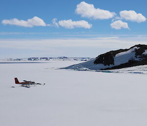 Flat expanse of sea ice with rocky hill mid picture to right, blue sky with white cumulous clouds above horizen. Cetre left of picture, red and white plane on the ice.