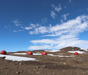 Rocky rolling hills to horizen at mid picture. Blue skies with 3 octas of white stratus cloud. Mid picture four huts which are round red melons across the landscape.