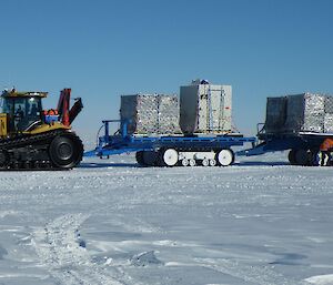 Snow covered ground, stretching to distance. Blue sky above horizen. Mid picture a yellow tractor pulling two blue sled carrying large pallets of netted cargo