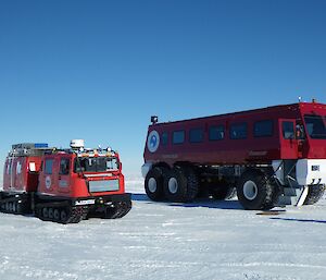 Snow covered ground stretching to horizen, above is bright blue sky. Mid picture right is a large red bus with whie steps from front door and white bull bar. To right a small red hagglunds vehicle, approximately the third of the size of the terrabus