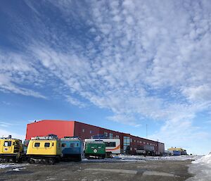 Top two thirds of picture is large expance of blue sky with cirrocumulous clouds. Bottom third of picture, dirt ground then large red building with yellow, blue and green hagglunds parked in front to left