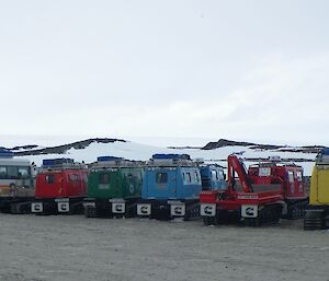 Line of vehicles mid picture, L to R — white and orange bus, red hagglunds, green hagglunds, blue hagglunds, red hagglunds with crane on trailer, then yellow hagglunds.