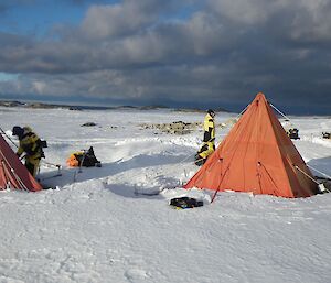 White snow covered ground to horizen, Blue sky peeking through grey clouds. Mid picture — two orange pyramid shaped tents with three expeditioners moving around them.